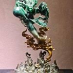 This jade sculpture depicts the mythical Gu Diao（蛊雕）. With horns, fierce fangs, sweeping wings, and a gripped orb, it conveys majestic power. Rich emerald and translucent jade highlight intricate details. Perched atop golden clouds and pale jade peaks, the mythic predator transforms into a guardian, symbolizing strength, protection, and soaring fortune.